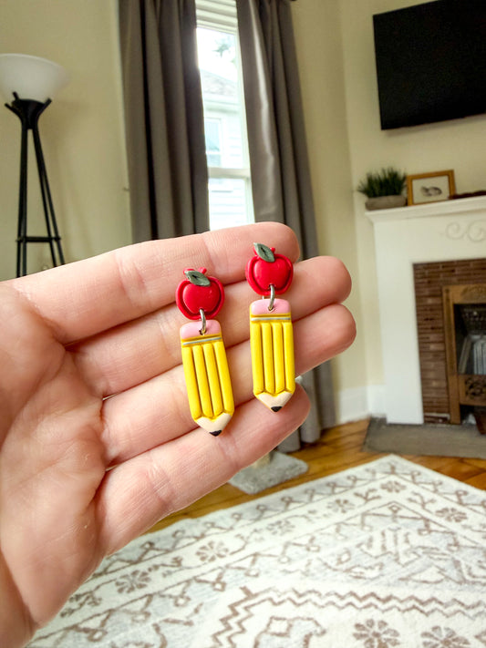 A hand holds a pair of novelty earrings shaped like yellow pencils with red apples on top, shown in a real indoor room with a window, curtains, fireplace, and rug in the background.