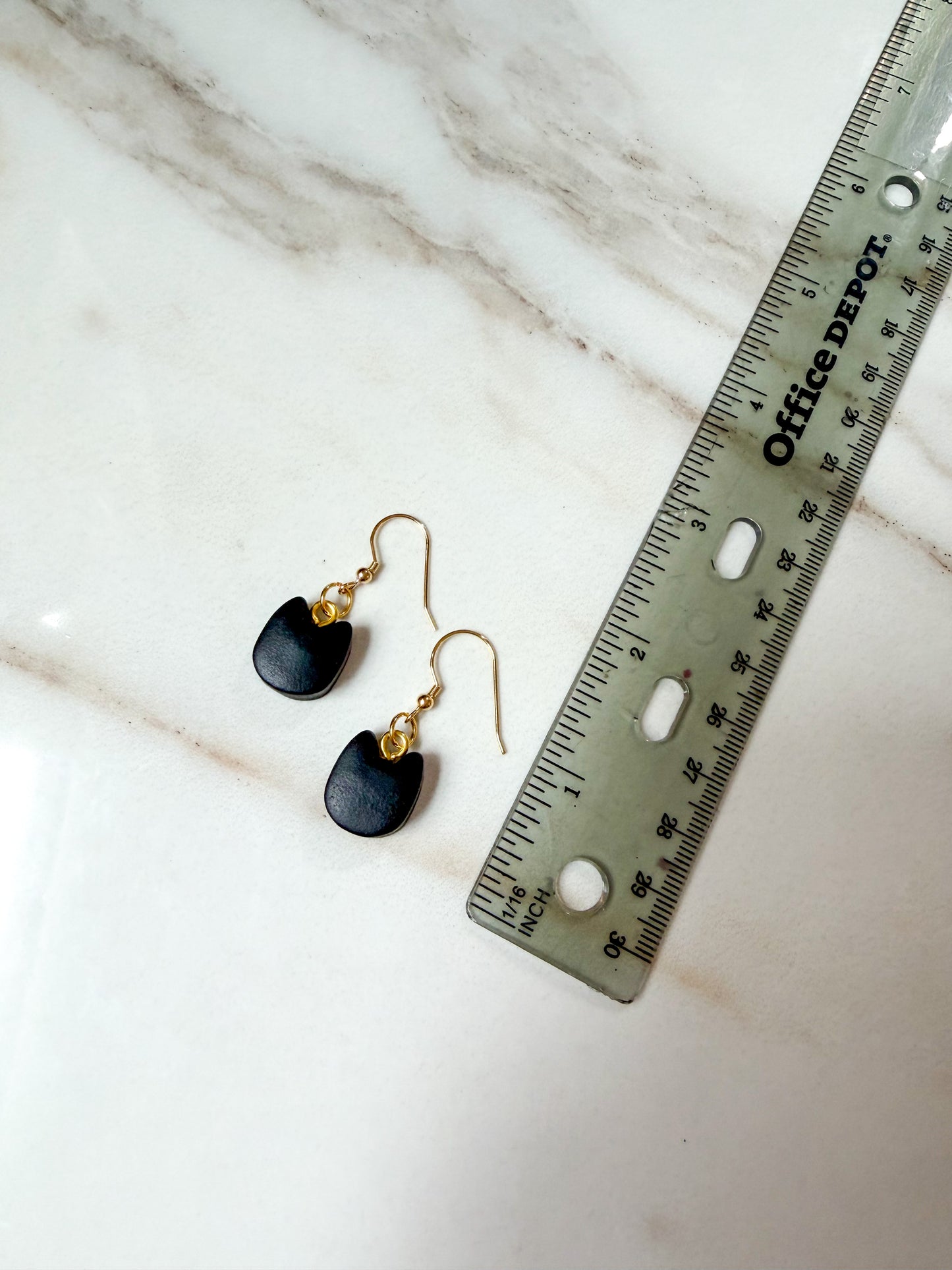 A pair of small black cat head-shaped drop earrings with gold fishhook wires, displayed next to a metal ruler on a white marble surface. Just under 2 inches in length. 