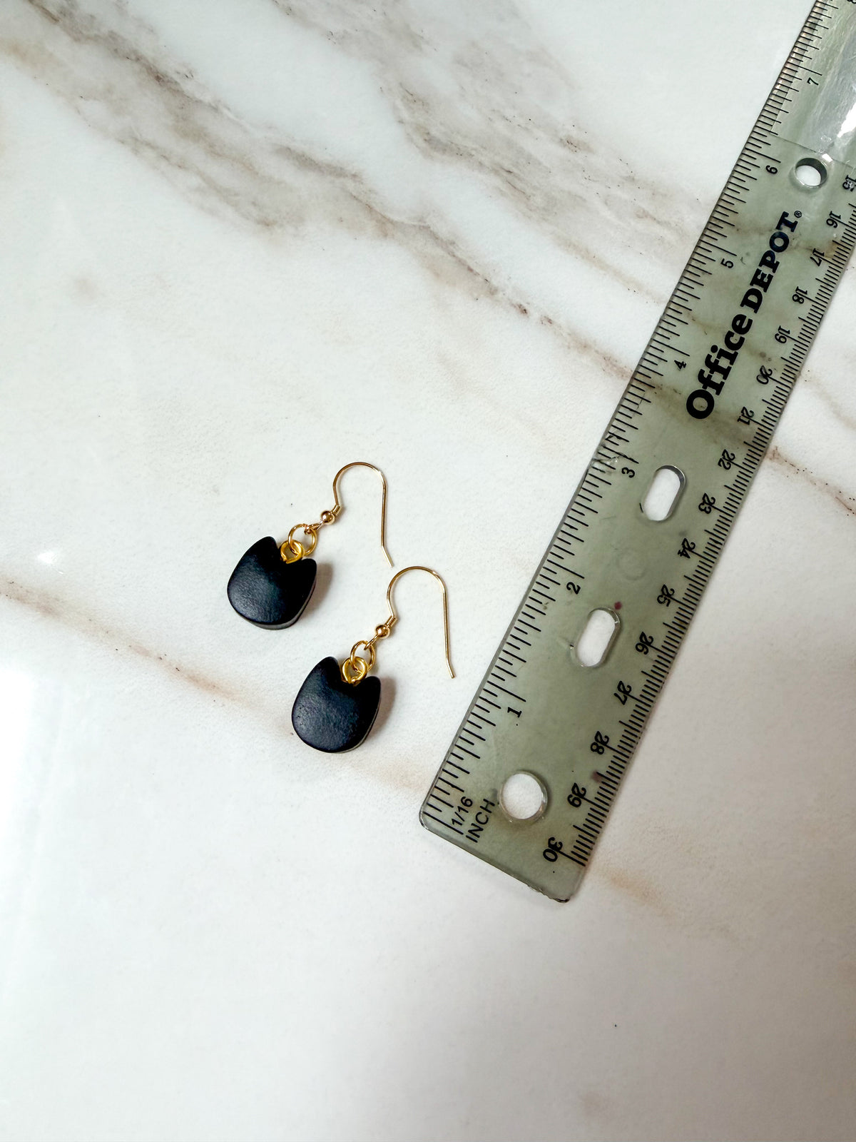 A pair of small black cat head-shaped drop earrings with gold fishhook wires, displayed next to a metal ruler on a white marble surface. Just under 2 inches in length. 