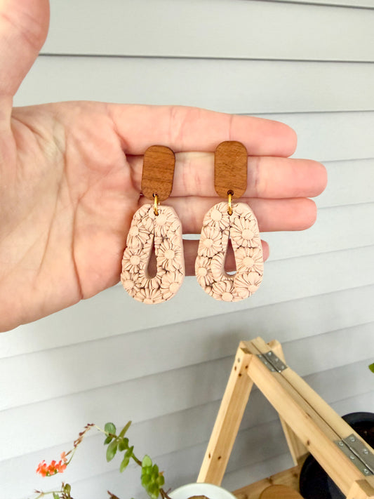 Tan colored earrings held in a hand. The earrings have textured flowers covering the surface. 