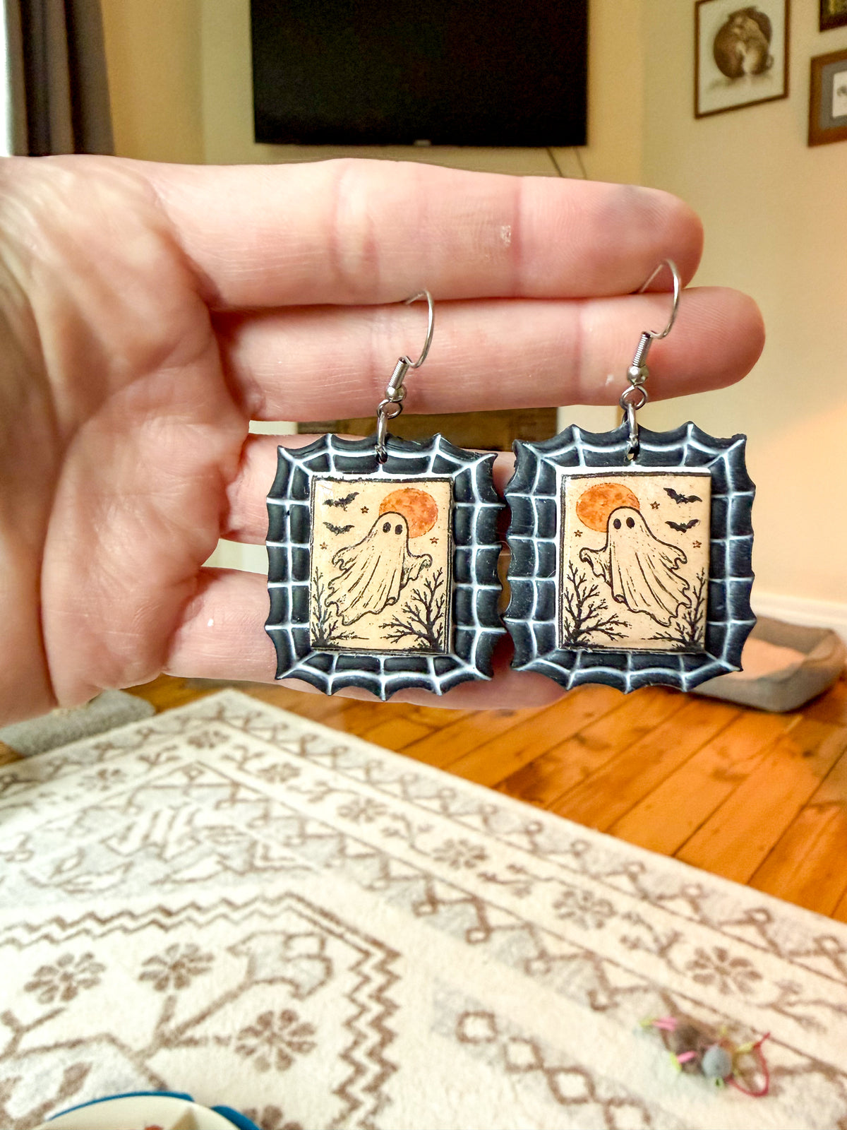 A hand holds a pair of Halloween-themed earrings with spiderweb borders and ghost illustrations in a home setting.