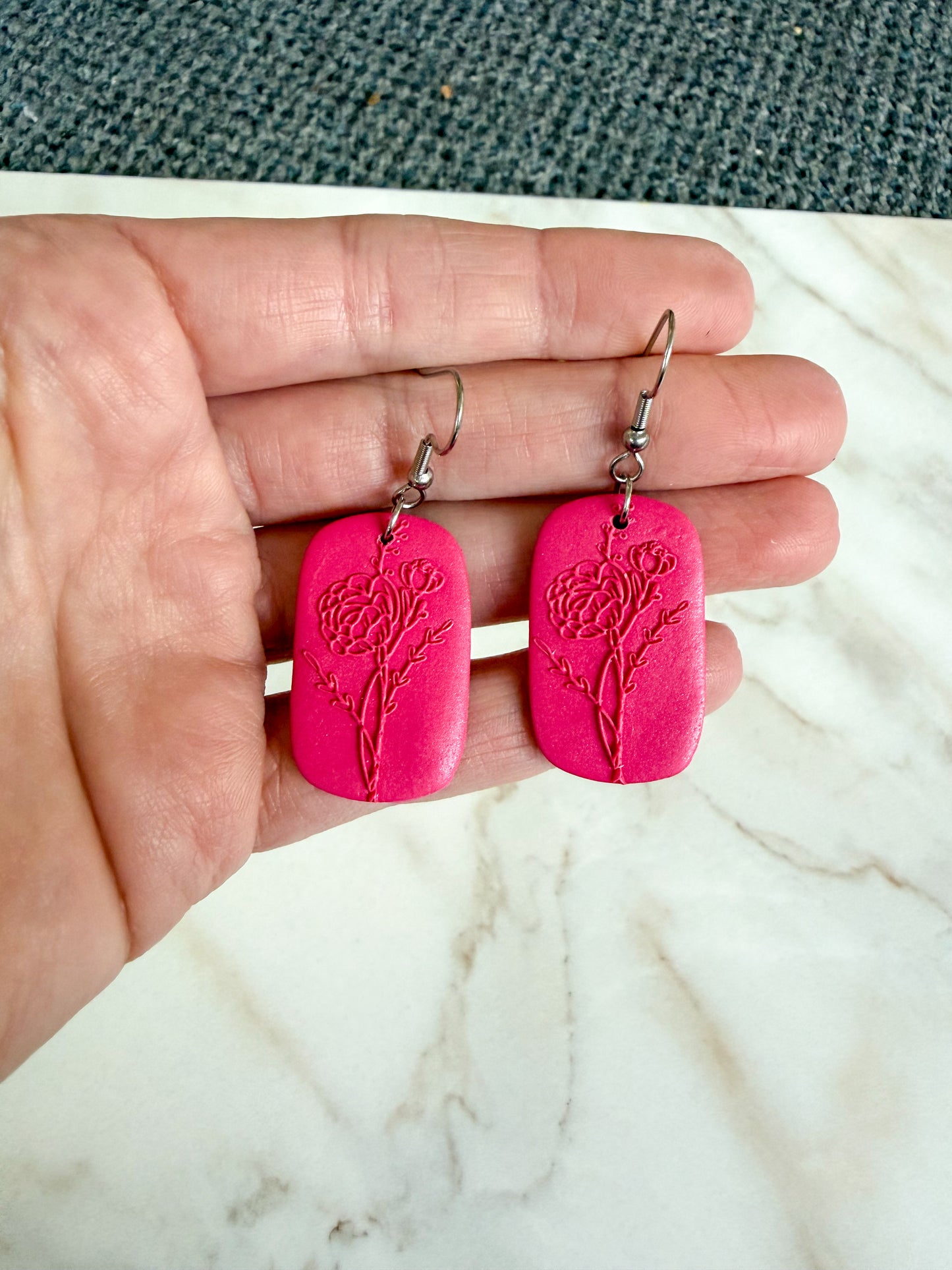 Pink earrings with floral design held in a hand on a marble surface