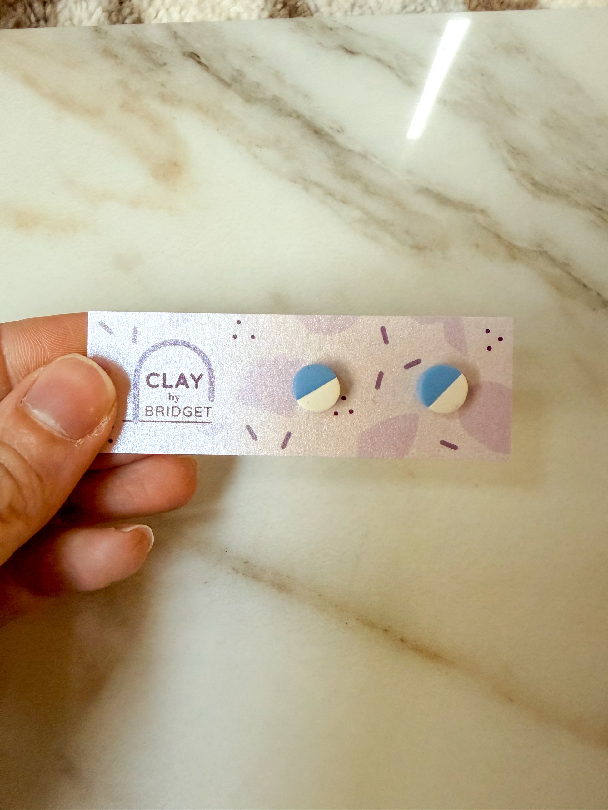 A hand holding a purple earring card with circular blue and white studs. 