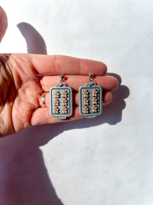 Pair of earrings held between fingers against a white background. The earrings are silver trays with tiny gingerbread men on them. 