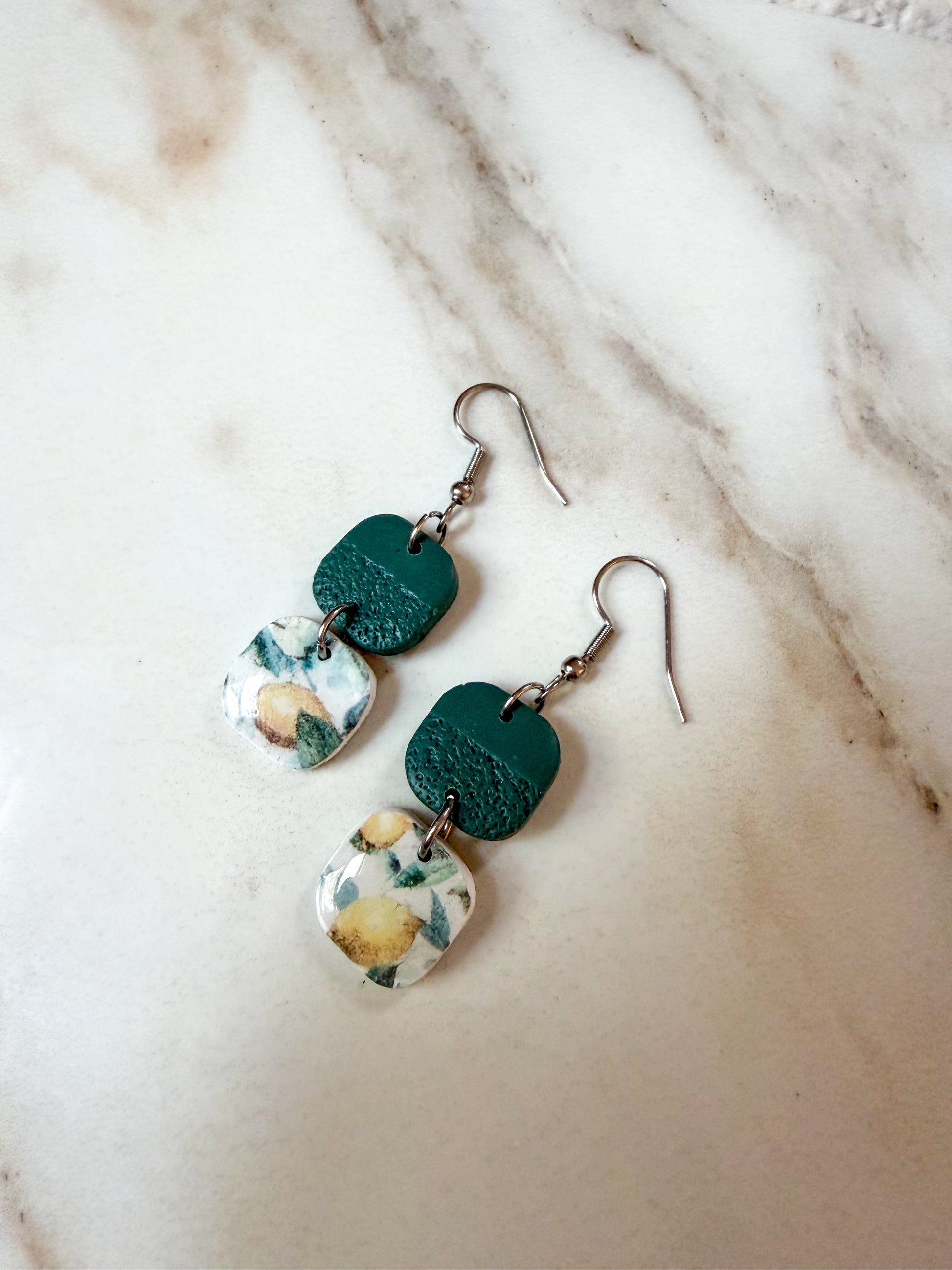 A pair of handmade dangle earrings with textured dark green charms and lemon patterned bottom charms displayed on a light marble surface.
