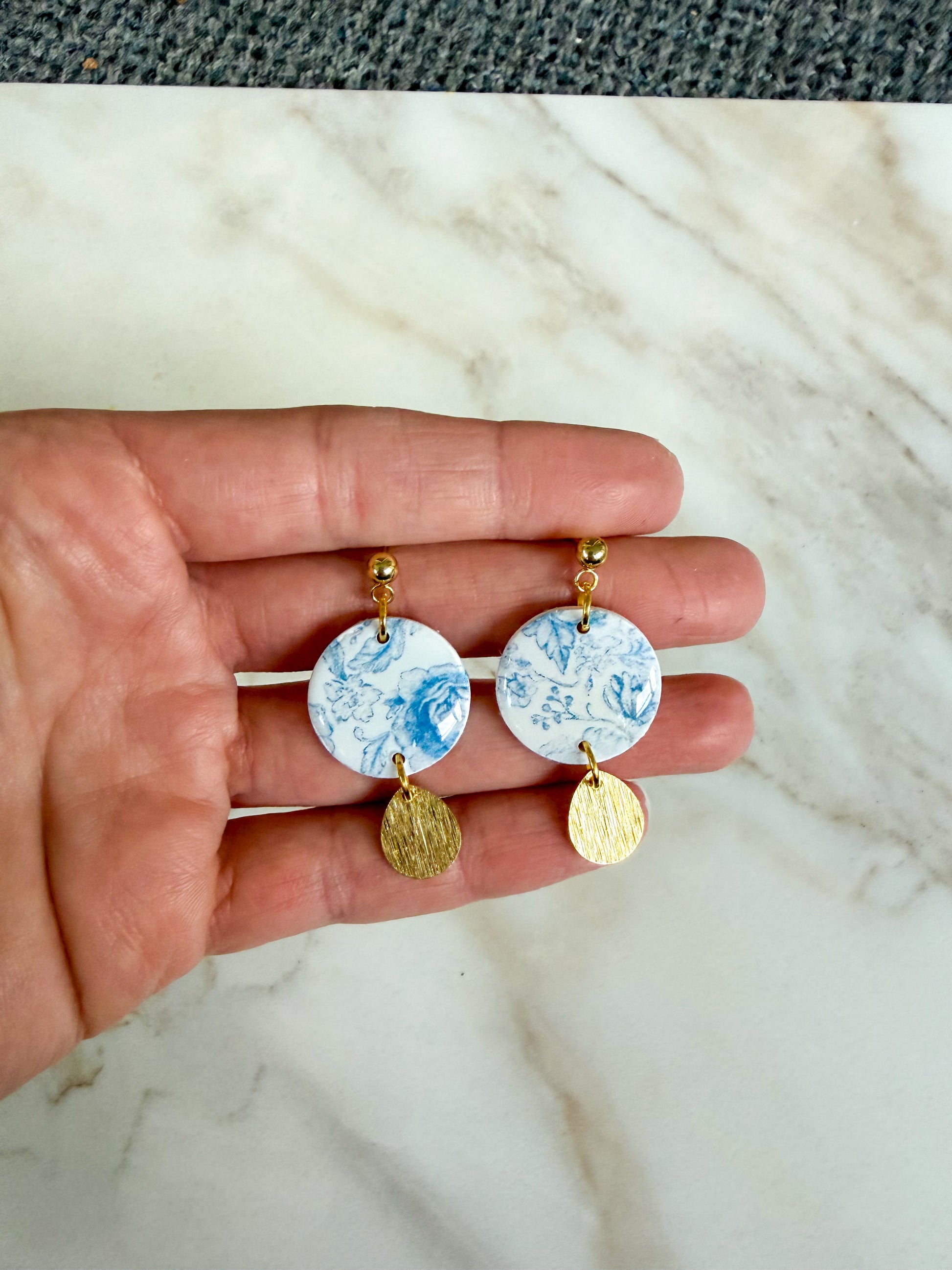 Hand holding two earrings with a blue floral design and gold accents on a marble background
