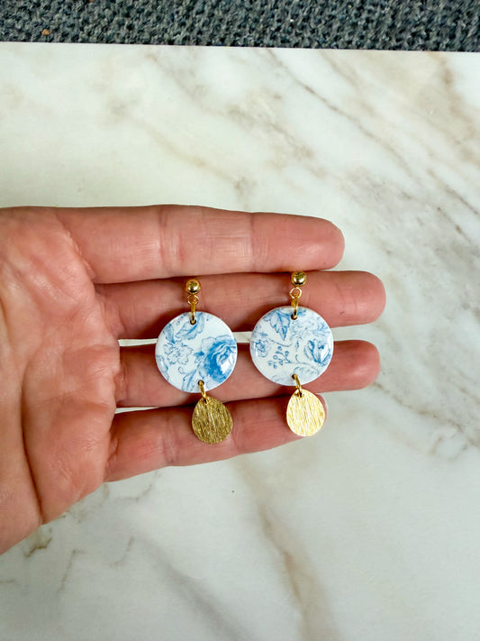 Hand holding two earrings with a blue floral design and gold accents on a marble background