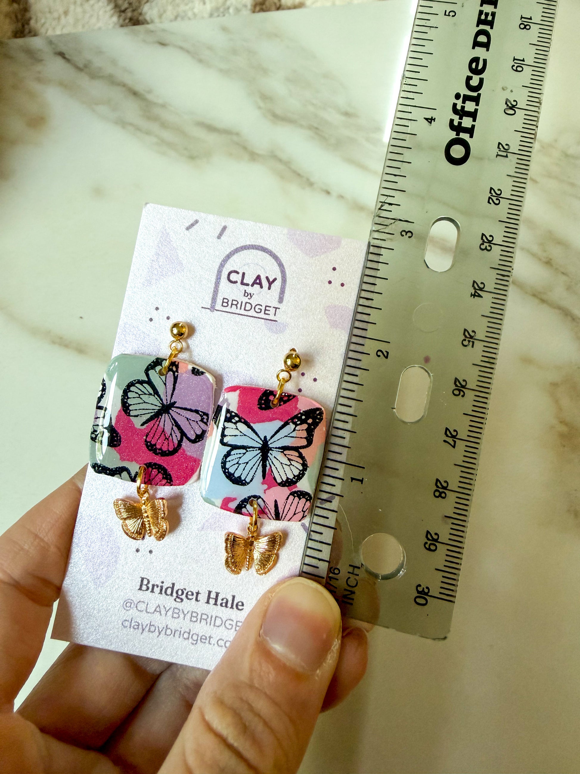 A hand holds a display card showing a pair of butterfly earrings next to a ruler for scale. The earrings have colorful butterfly-shaped charms with pink, purple, and blue patterns, gold-tone posts, and gold-tone butterfly drops. About 2 inches in length. 