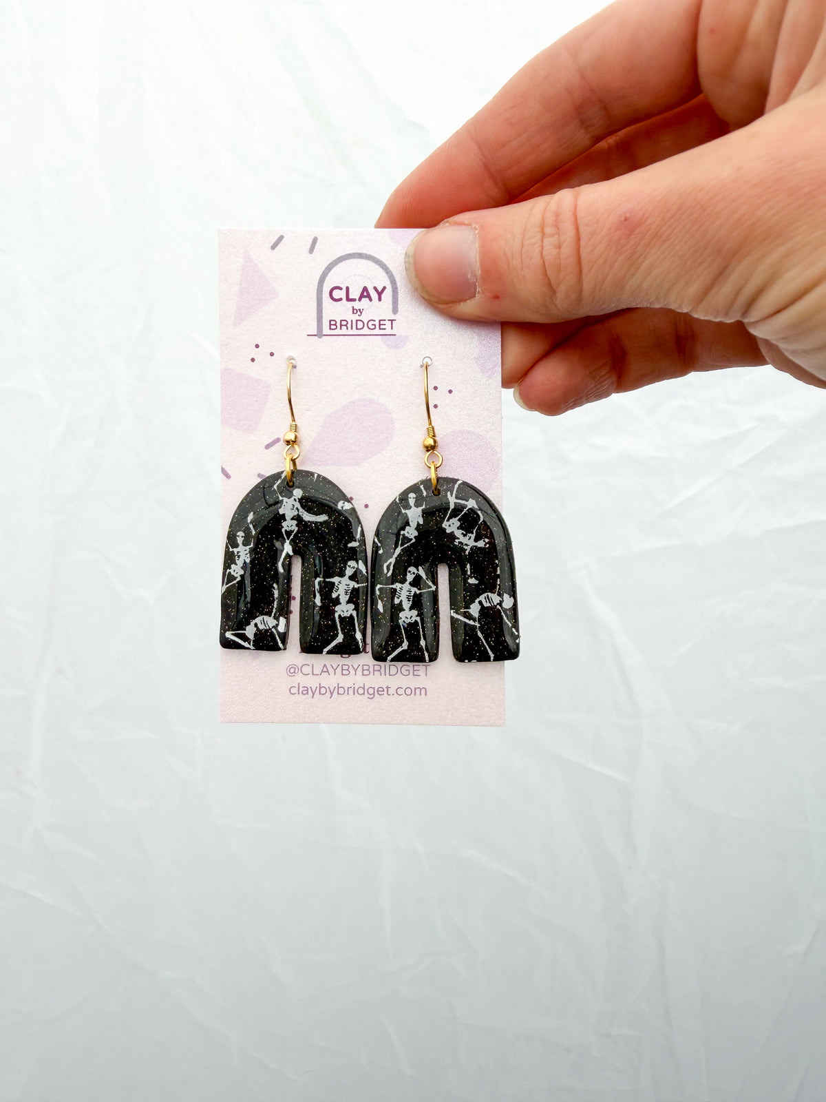 Glittery black arch shaped earrings with white skeleton designs. Held in the hand against the white surface.