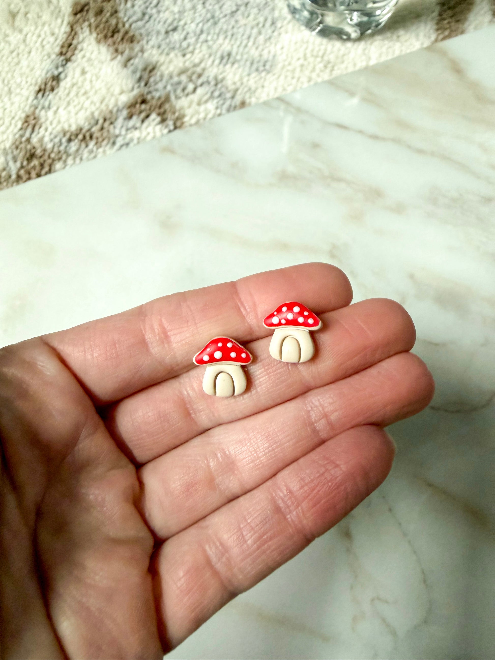 Two small mushroom-shaped earrings held between fingers on a marble surface.