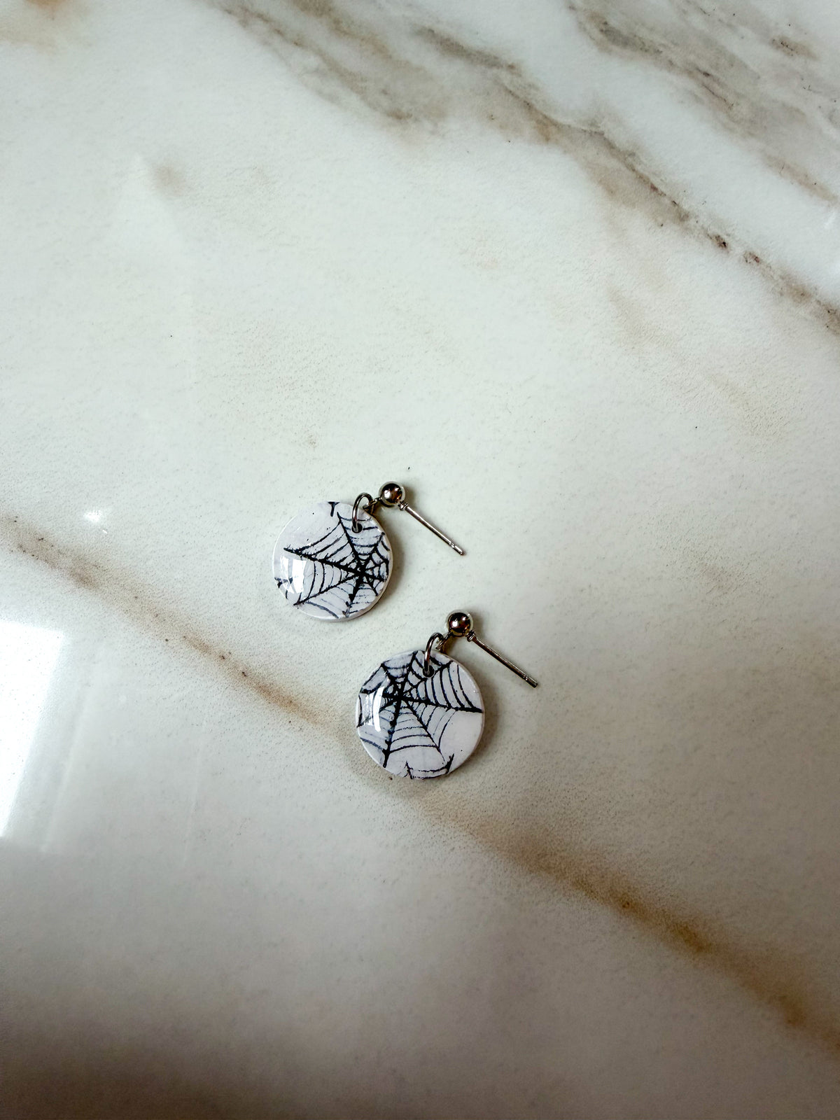 A pair of small round earrings with spiderweb designs on a white background, resting on a light marble surface.