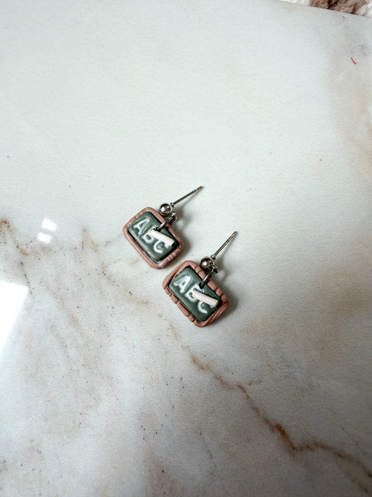 A pair of earrings on a marble surface. Made to resemble little chalkboards with a piece of chalk. It reads ABC. 