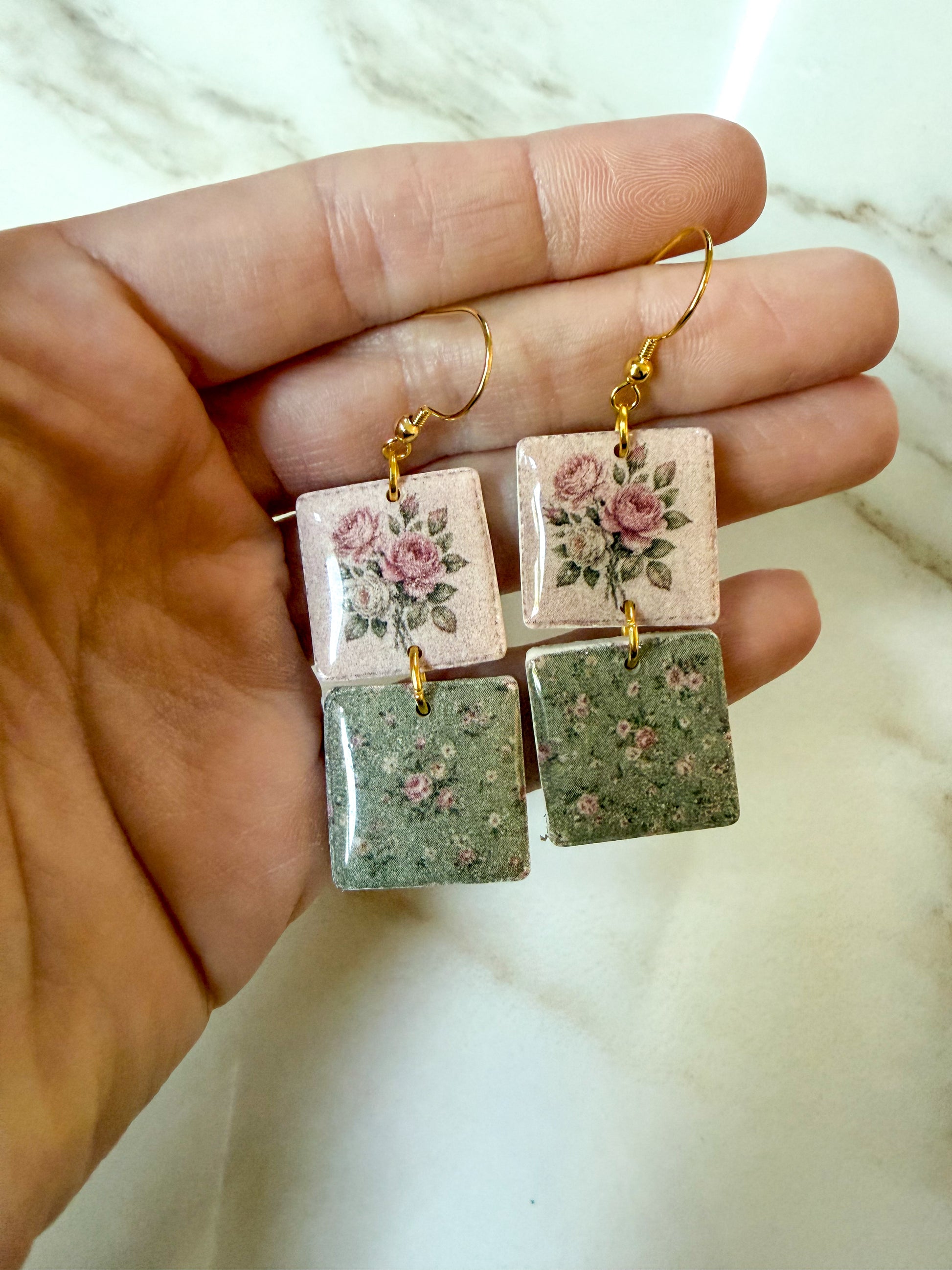 A pair of handmade square dangle earrings held in a hand. The earrings feature a top floral square with pink roses and green leaves on a light pink background, and a bottom textured square in sage green.