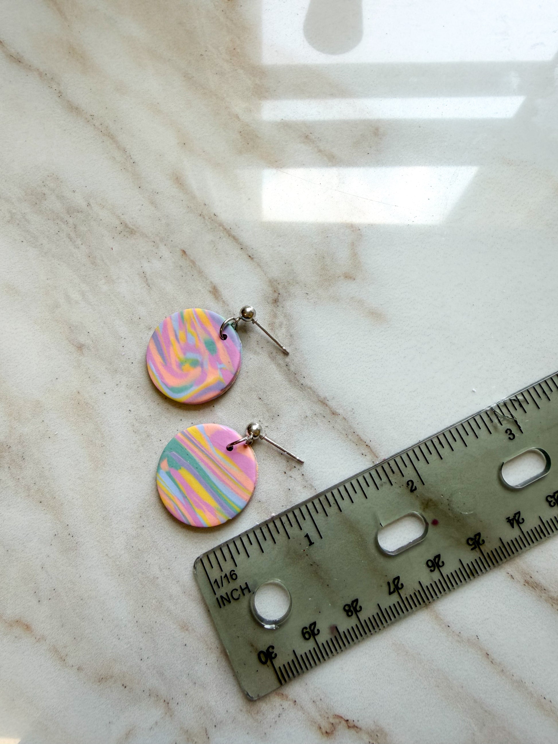 A pair of round dangle earrings with pastel pink, yellow, and blue marbled patterns on silver posts, shown next to a ruler on a light marble surface. About one inch in length.