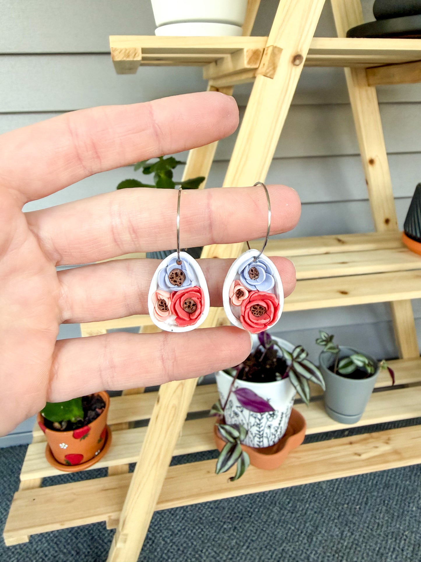 Hand holding two floral-themed earrings with a wooden shelf and plants in the background