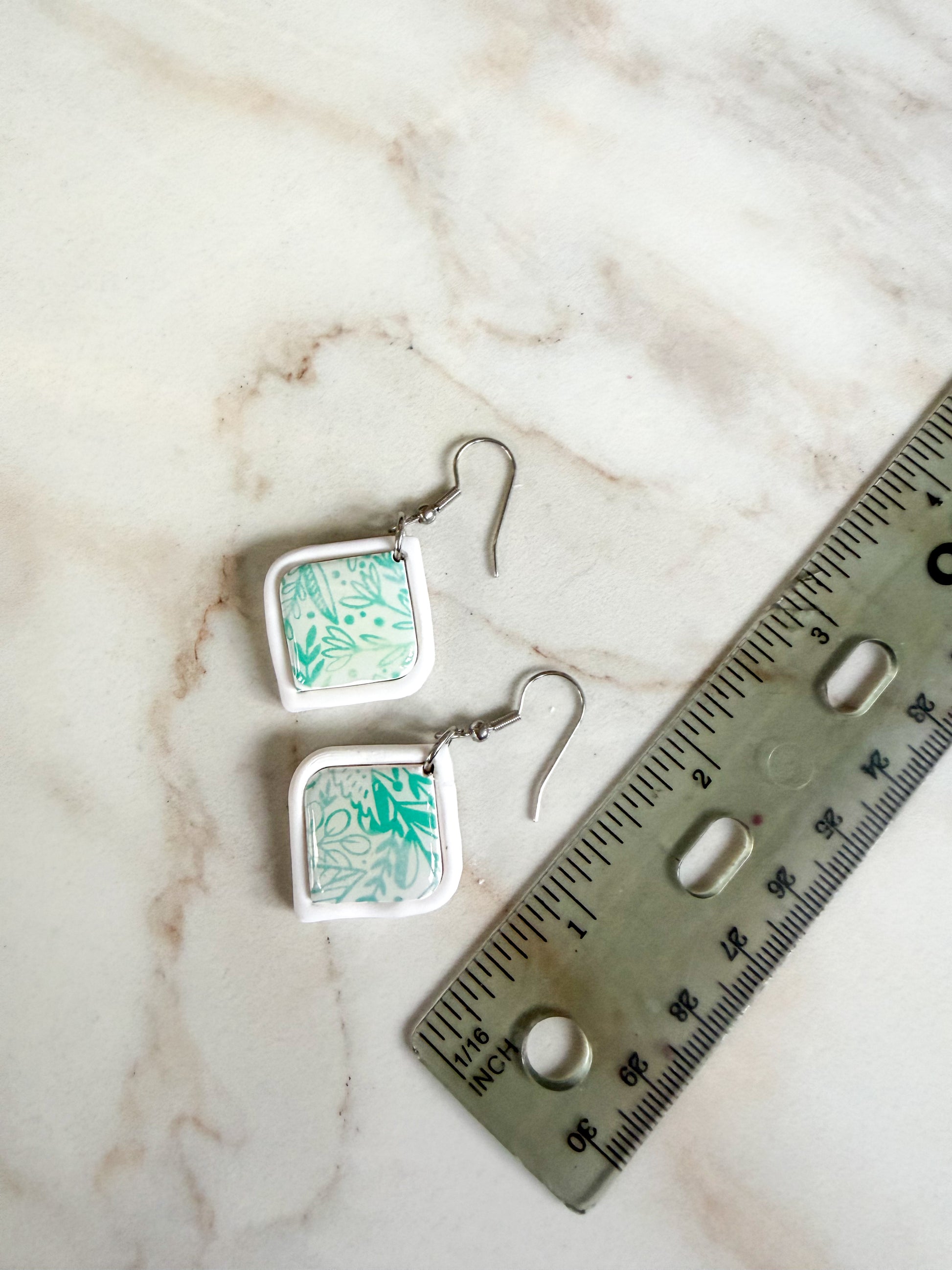 Green and white floral earrings lying on a marble surface next to a ruler for scale. About 2 inches in length.  