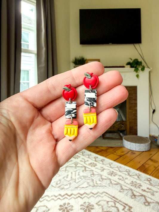 A hand holding a pair of colorful earrings shaped like red apples, black-and-white notebooks, and yellow pencils, with a blurred living room in the background.