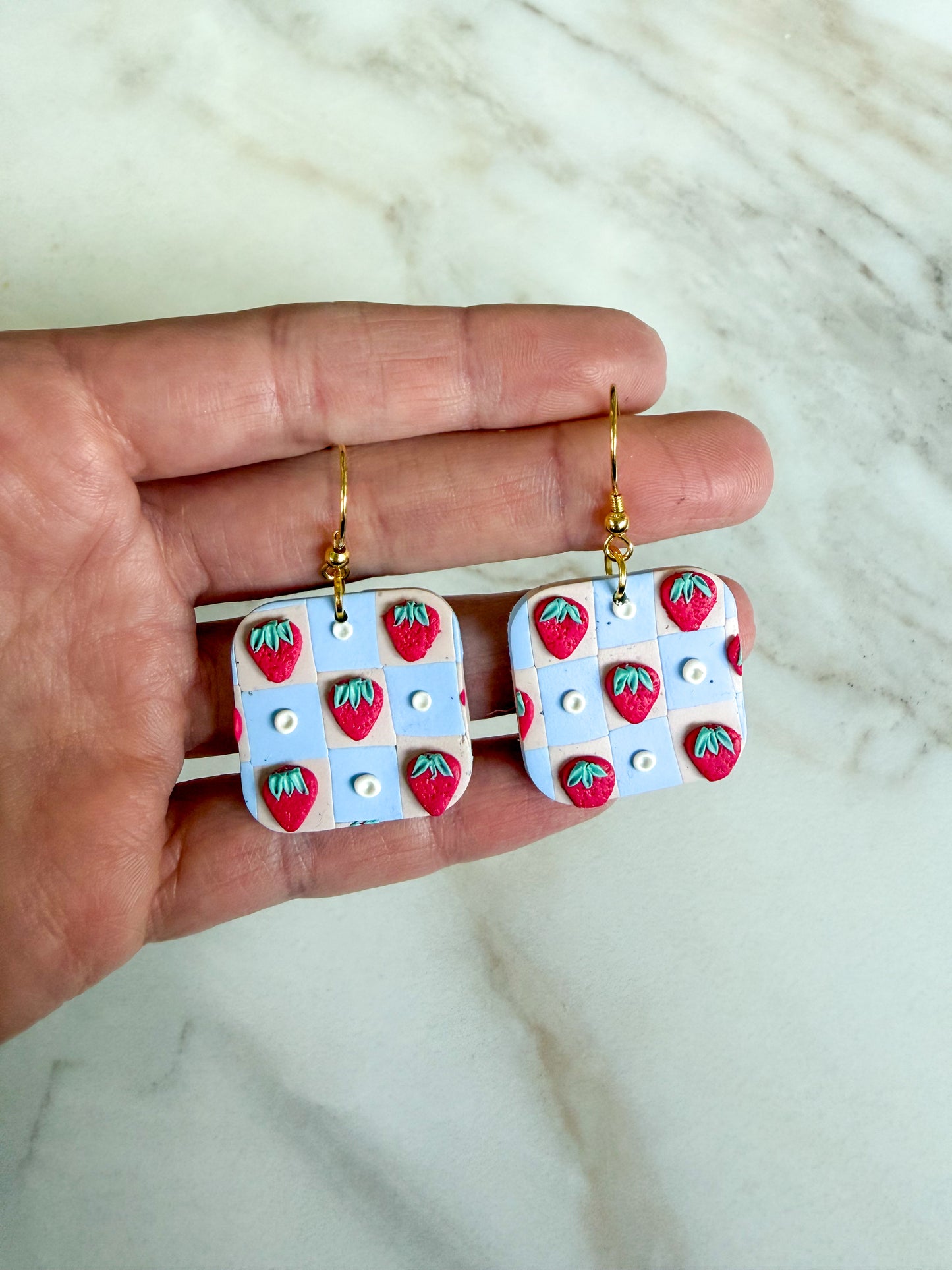 Earrings with strawberry pattern held between fingers on a marble background