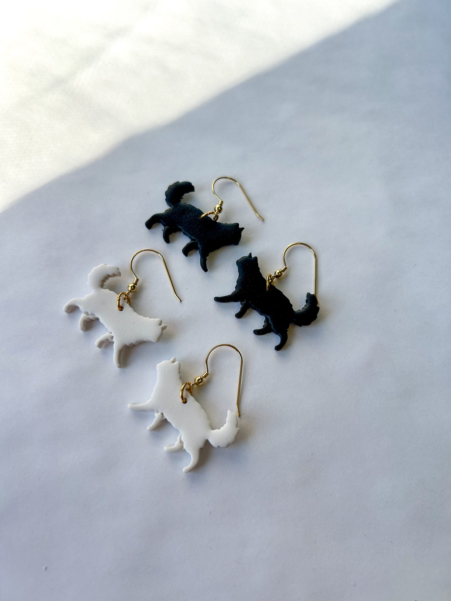 Cat-shaped earrings on a light gray surface. One pair is black and the other pair is white. Long-haired cats.
