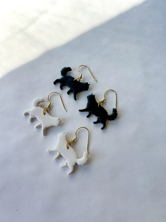 Cat-shaped earrings on a light gray surface. One pair is black and the other pair is white. Long-haired cats.