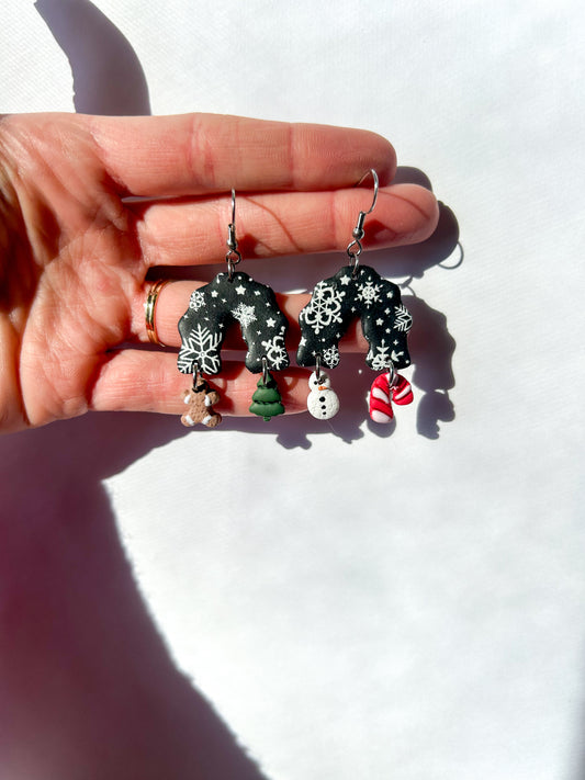 Hand holding Christmas-themed earrings against a white background. Black arch dangles with a snowflake design. The four charm hanging off of the earrings are a tree, a gingerbread man, a candy cane, and a snowman
