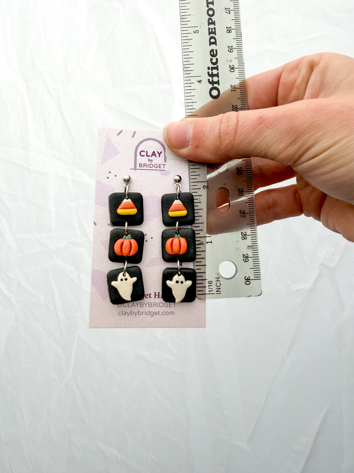 A hand holds a pair of Halloween-themed clay dangle earrings against a white background, with a ruler for scale. The earrings feature candy corn, pumpkins, and ghosts on a black square background. Around 2 inches in length. 