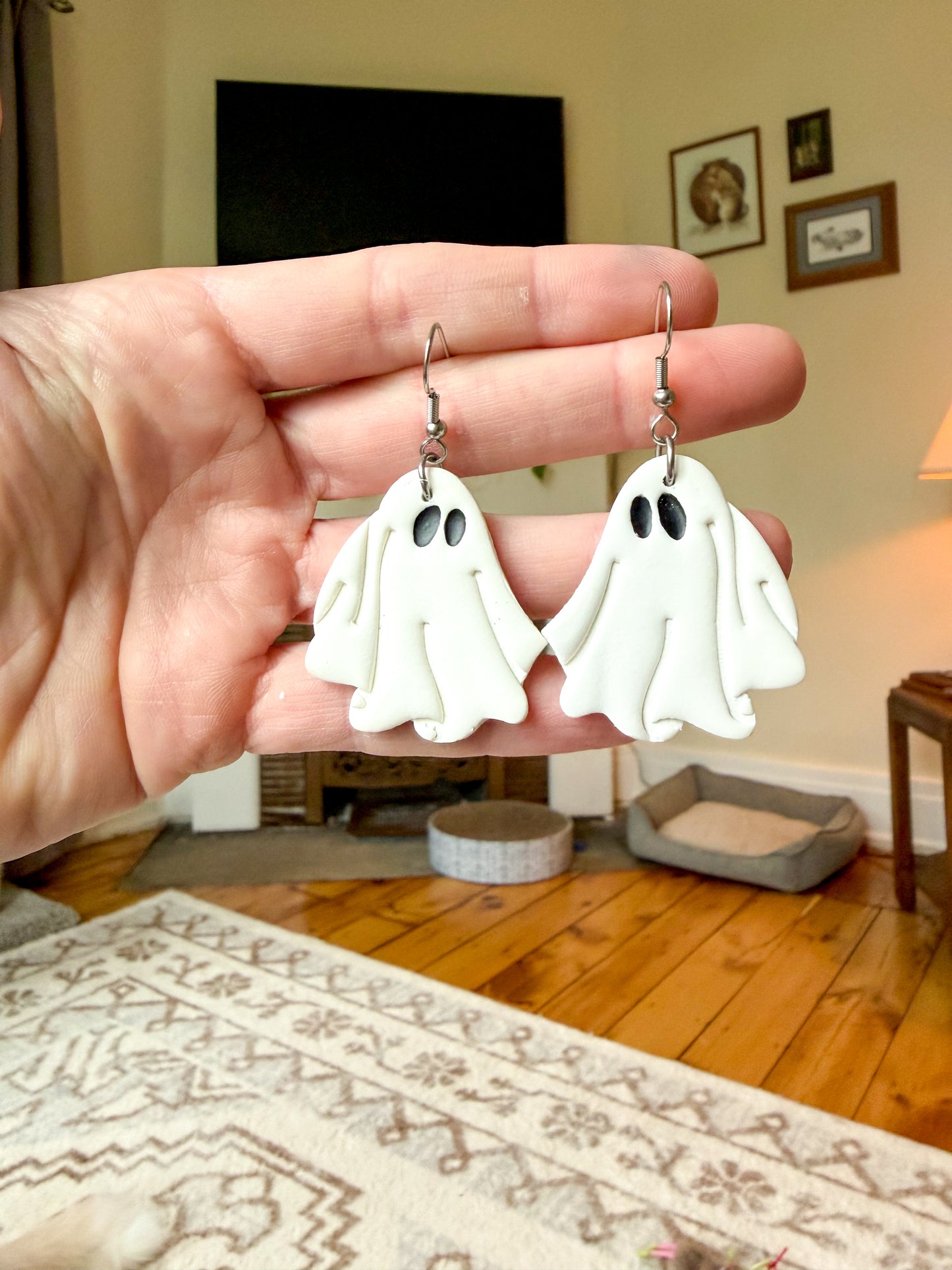 A hand holds a pair of white ghost-shaped dangle earrings with silver fishhook wires in a cozy, warm-toned living room setting.