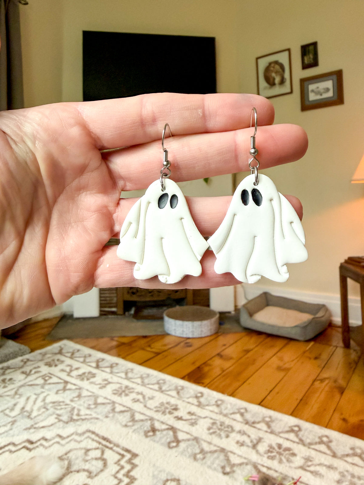 A hand holds a pair of white ghost-shaped dangle earrings with silver fishhook wires in a cozy, warm-toned living room setting.