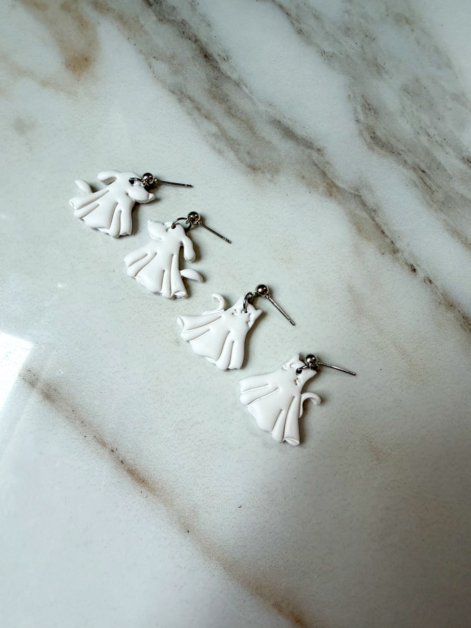 Four white cat and dog ghost earrings with silver posts, arranged diagonally on a white and gray marble surface.