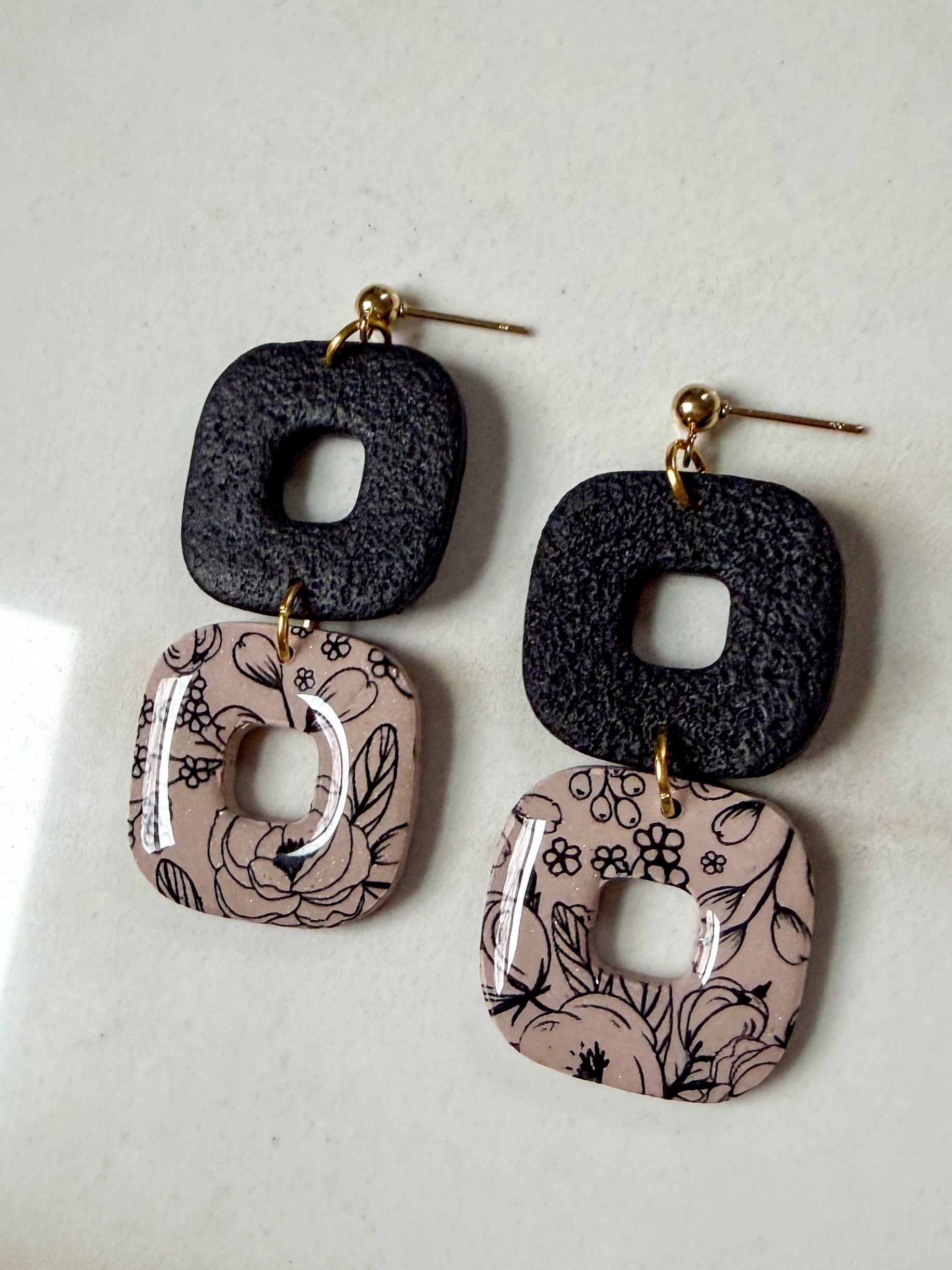 Square “donut” shape dangles with a black square on top and a shiny brown square on the bottom with a black floral design. 