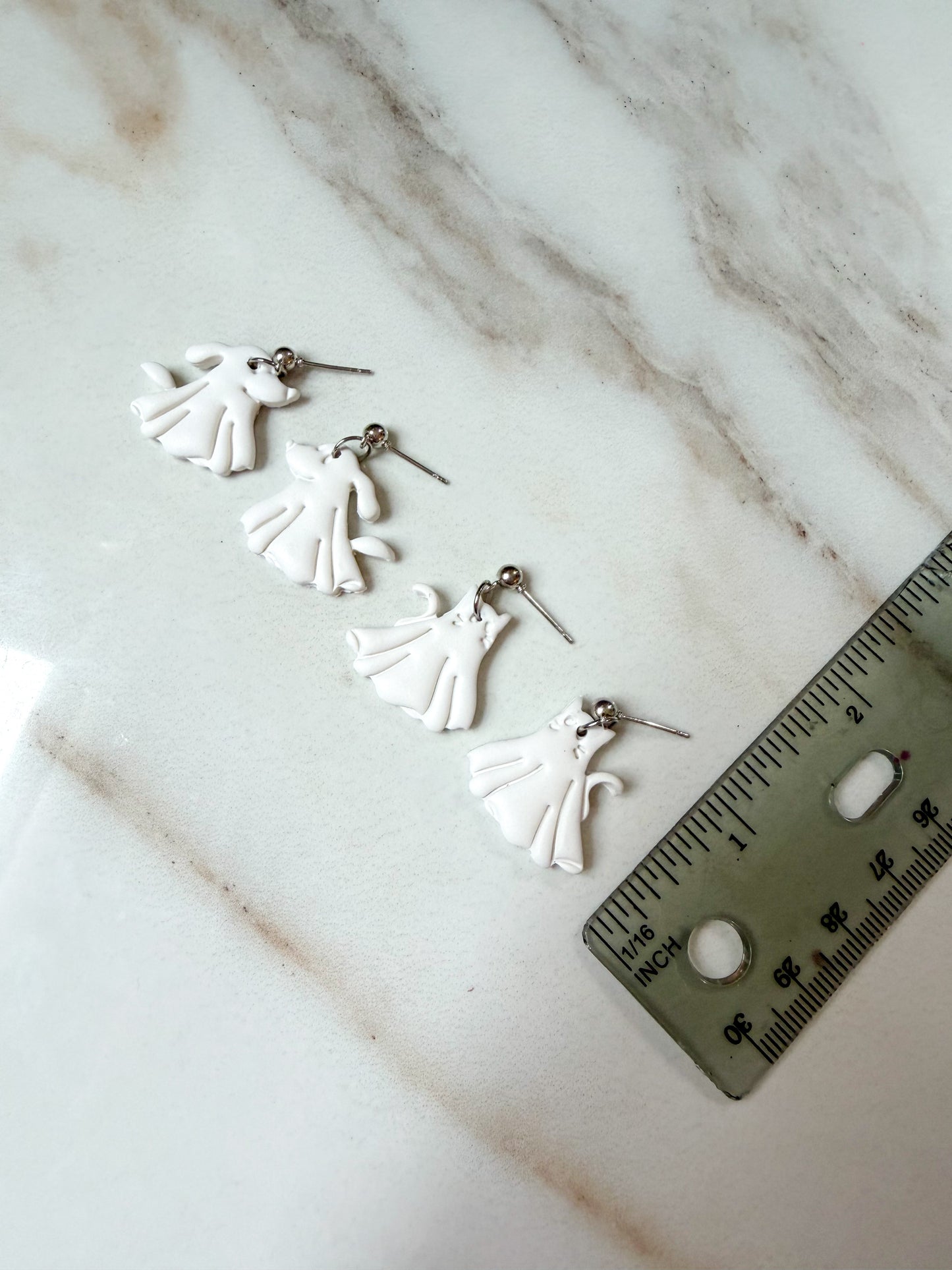 Four small white ghost-shaped earrings with silver posts are arranged on a white marble surface next to a metal ruler for scale. About 1 inch in length. 