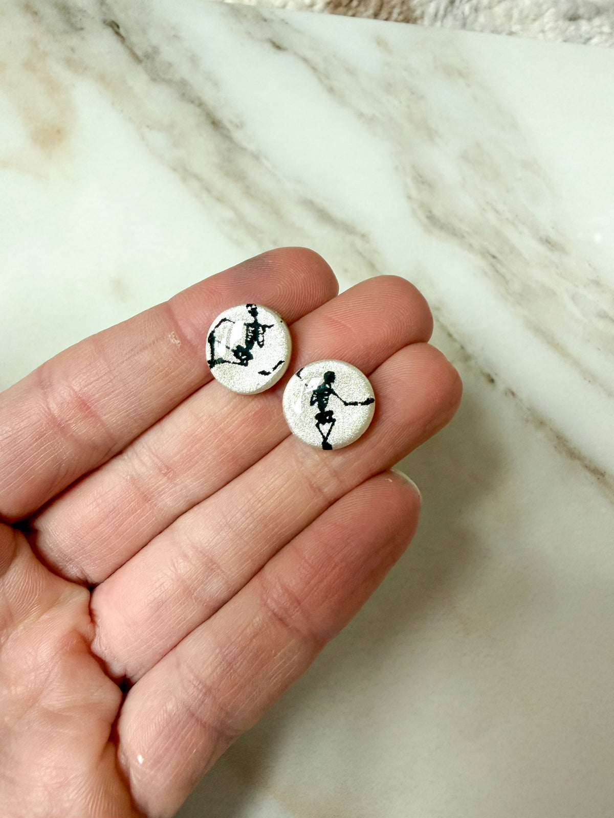A close-up of a hand holding two small round stud earrings with black skeleton figures on a white background, set against a light marble surface.
