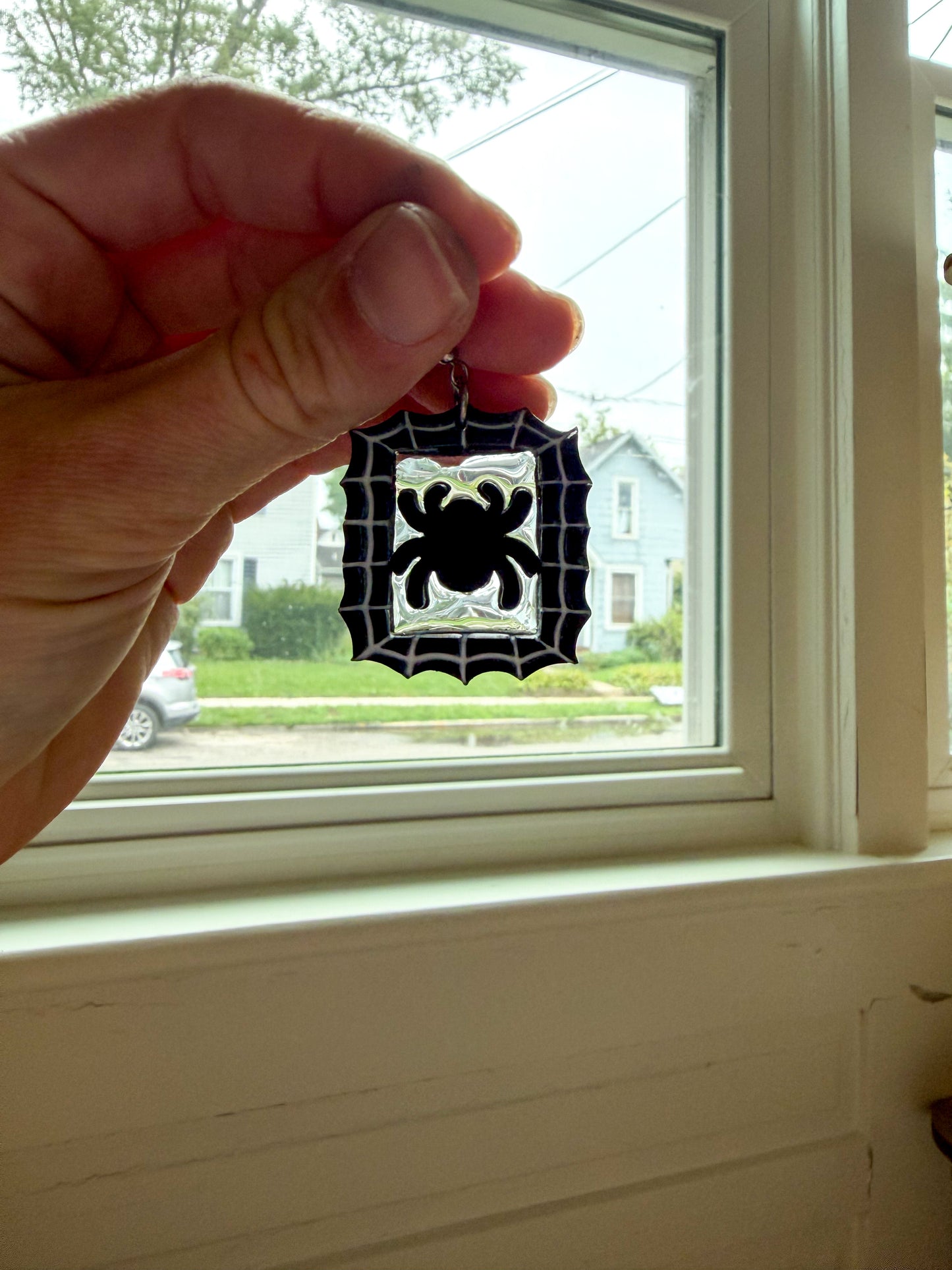 A hand holds a black spider-shaped earring in front of a window, with a house and yard visible outside.