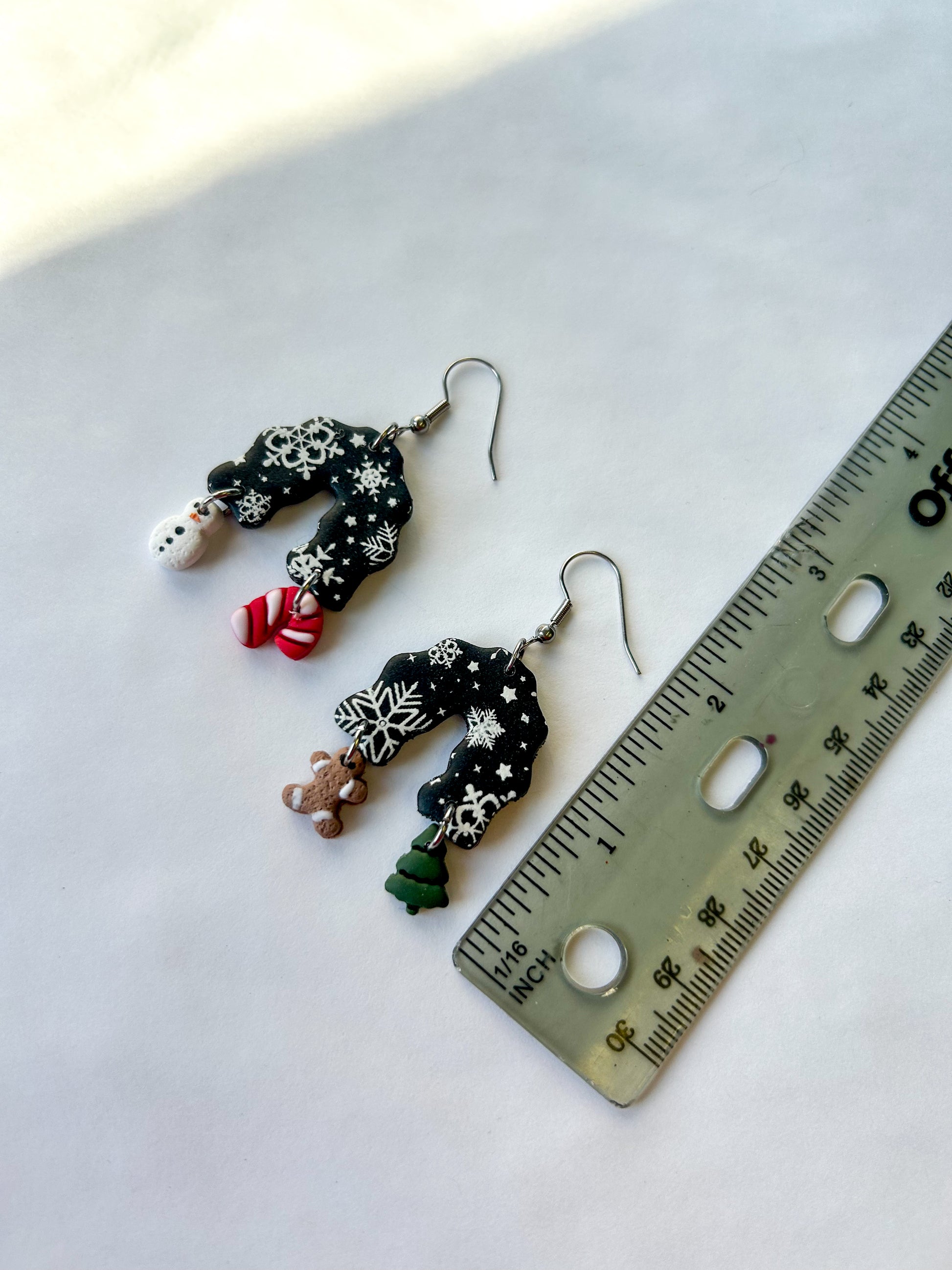 Pair of earrings next to a ruler on a light background. Black arch dangles with a snowflake design. The four charm hanging off of the earrings are a tree, a gingerbread man, a candy cane, and a snowman