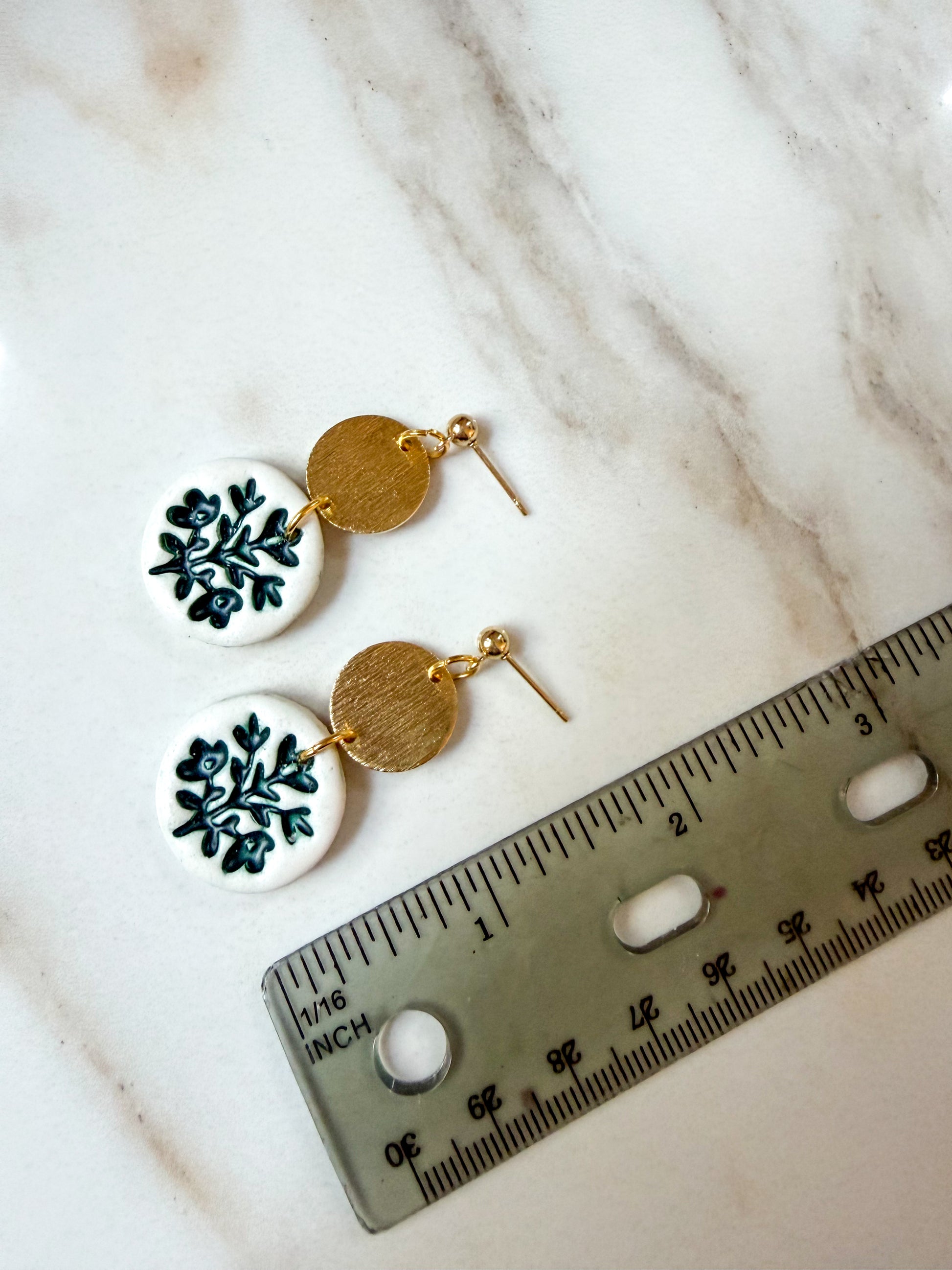 A pair of handmade dangle earrings with white circular discs featuring a green floral pattern, a textured gold disc, and gold fishhook wires, displayed next to a ruler on a white marble surface. About 2 inches long. 