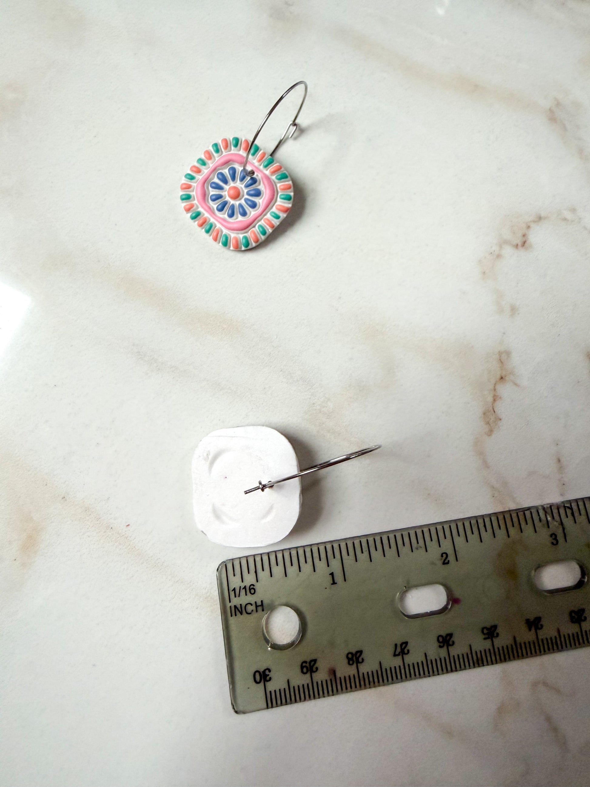 Colorful polymer clay earring on a marble surface with a ruler for scale. About an inch and a half long. 