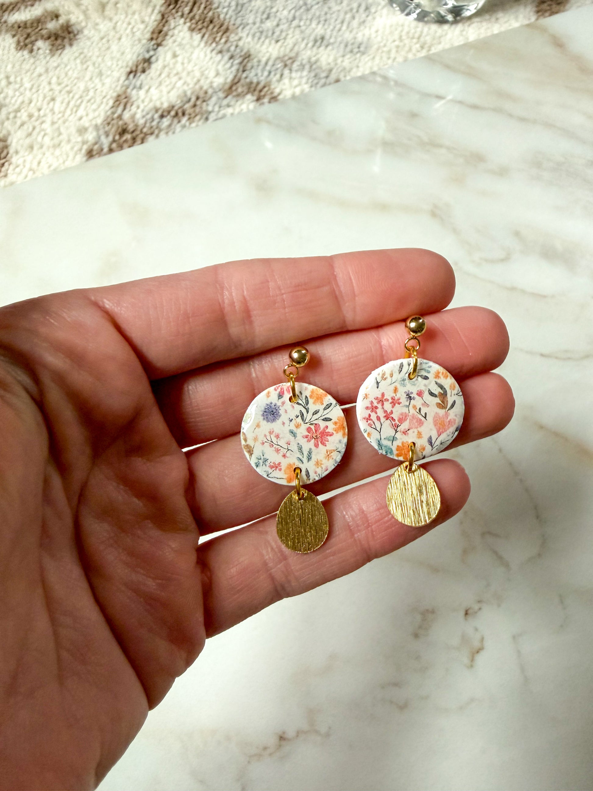 Hand holding a pair of floral earrings with gold accents on a marble surface.