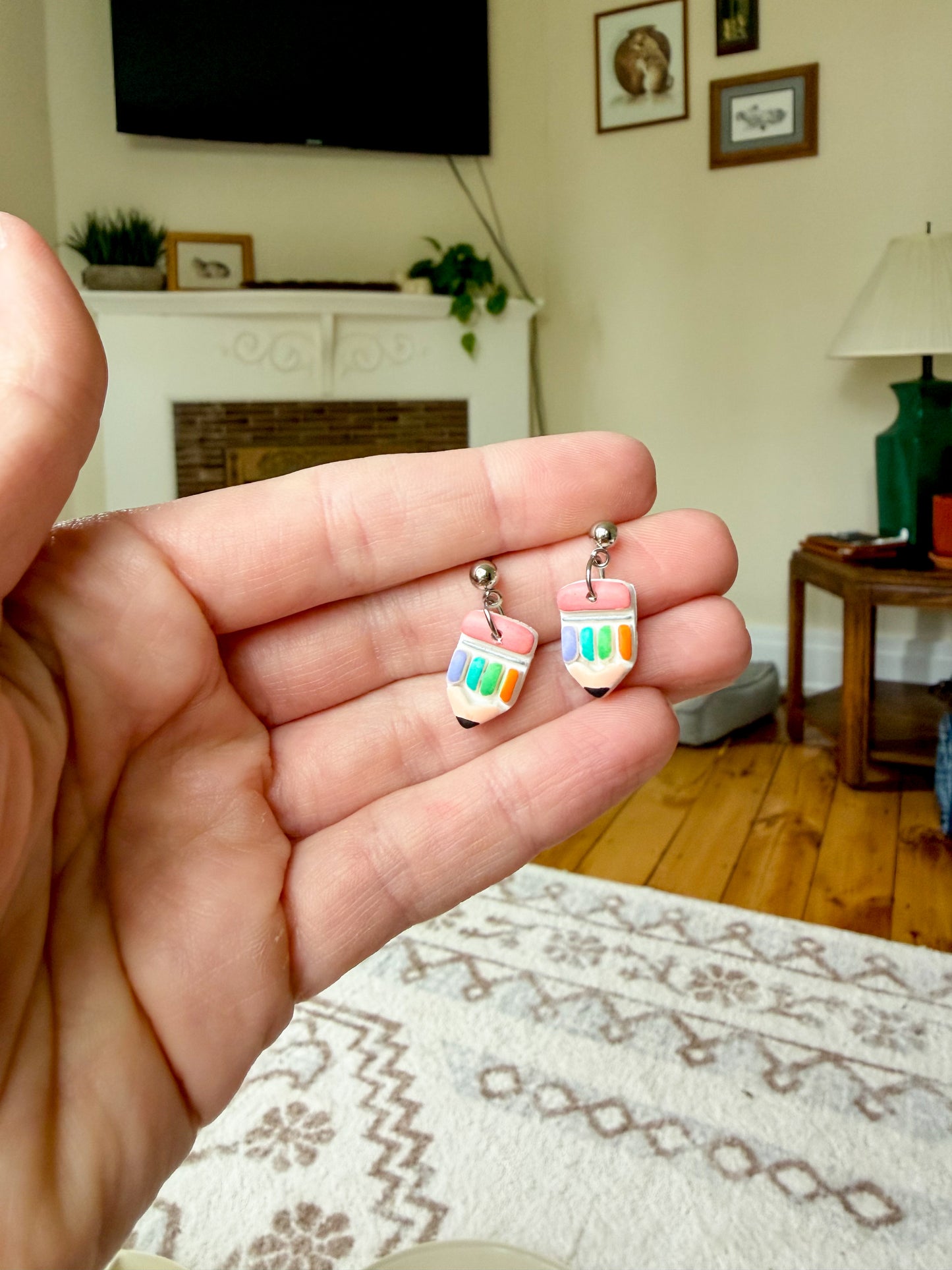 A hand holding a pair of small pencil-shaped earrings with colorful striped bodies and silver posts, photographed in a real living room setting.