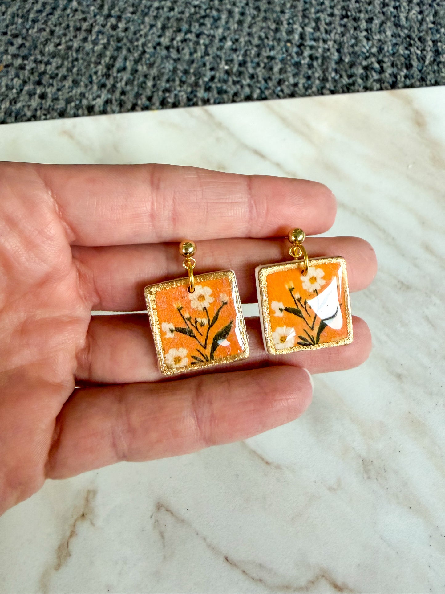 Hand holding a pair of square earrings with floral design on an orange background.