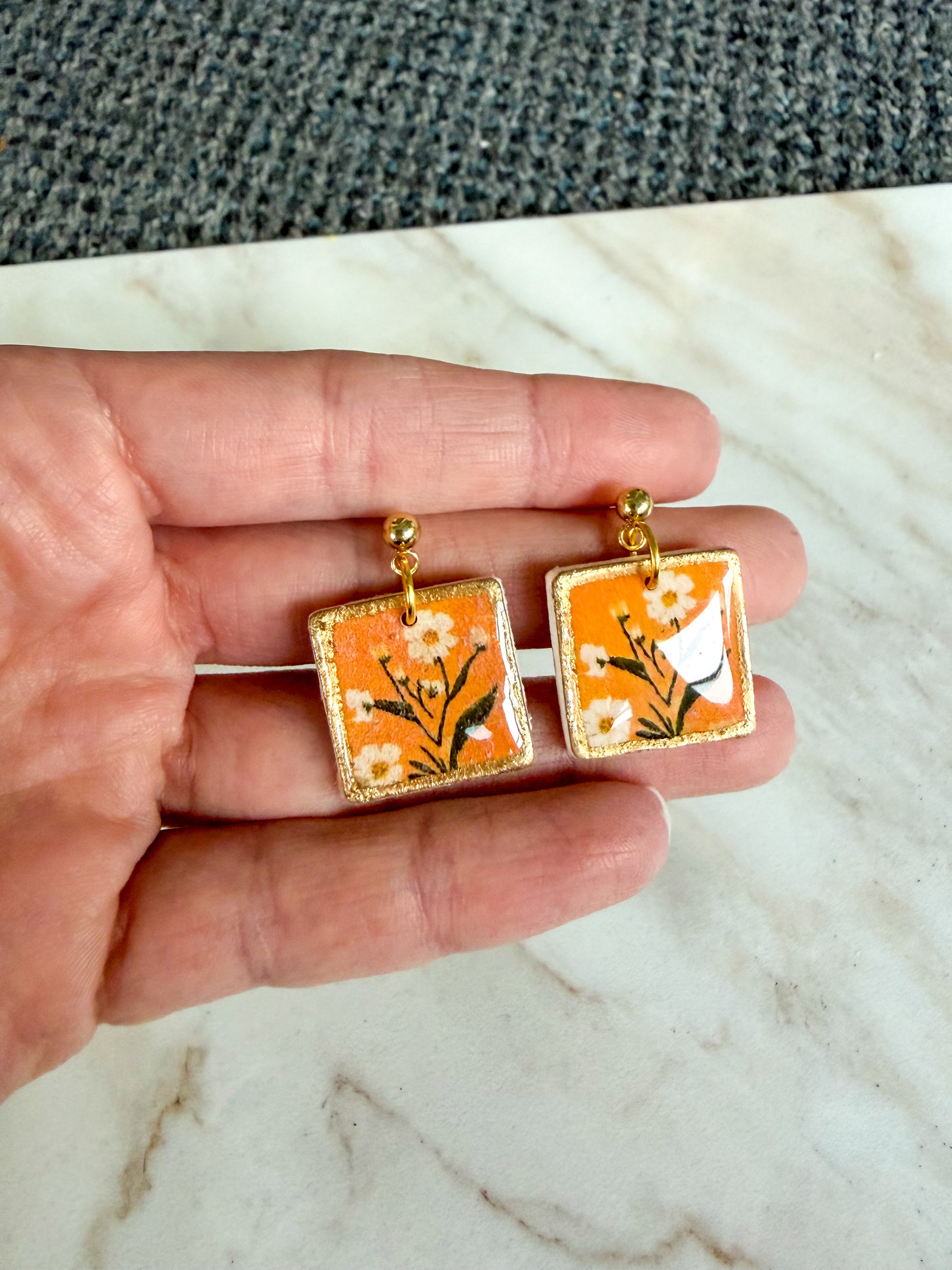 Hand holding a pair of square earrings with floral design on an orange background.