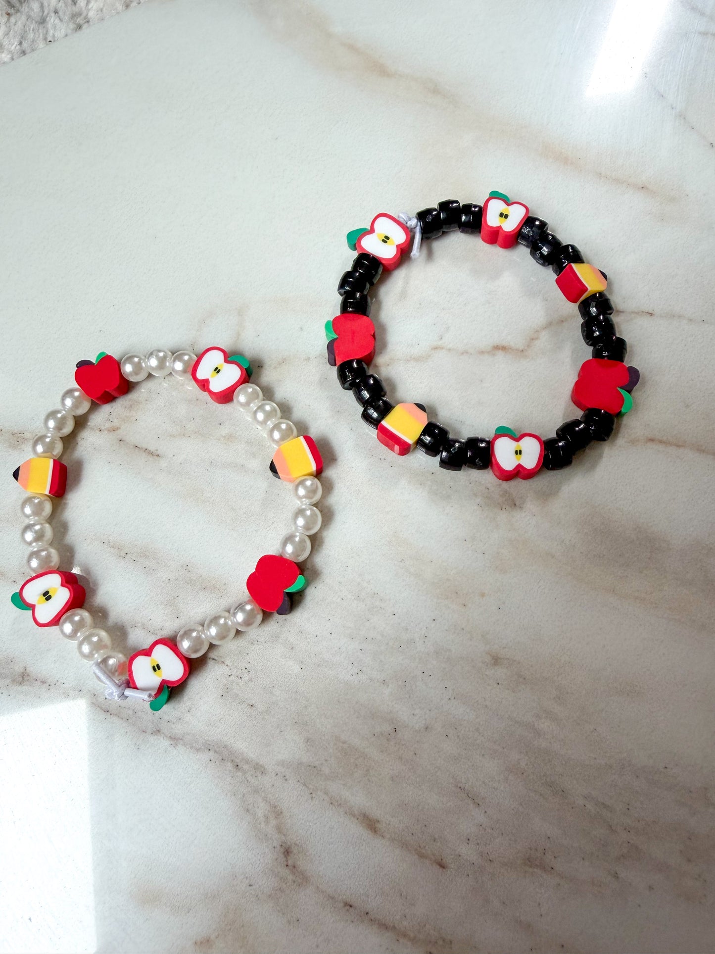 Two beaded stretch bracelets displayed on a light marble surface. One bracelet features white faux pearls with apple and pencil shaped beads. The other has black beads with the same apple and pencil beads. 