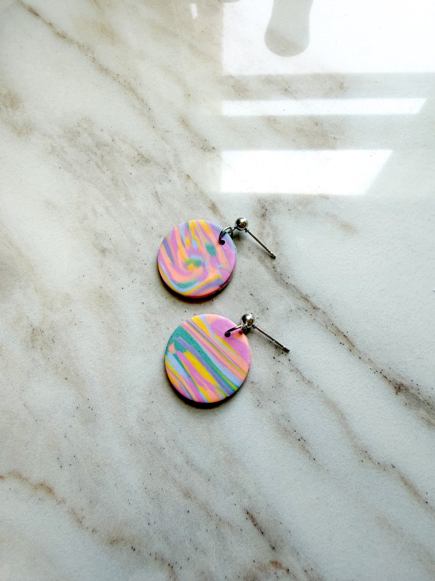 A pair of round dangle earrings with a pastel marbled pattern in pink, yellow, blue, and green, hanging from silver posts on a white marble surface.