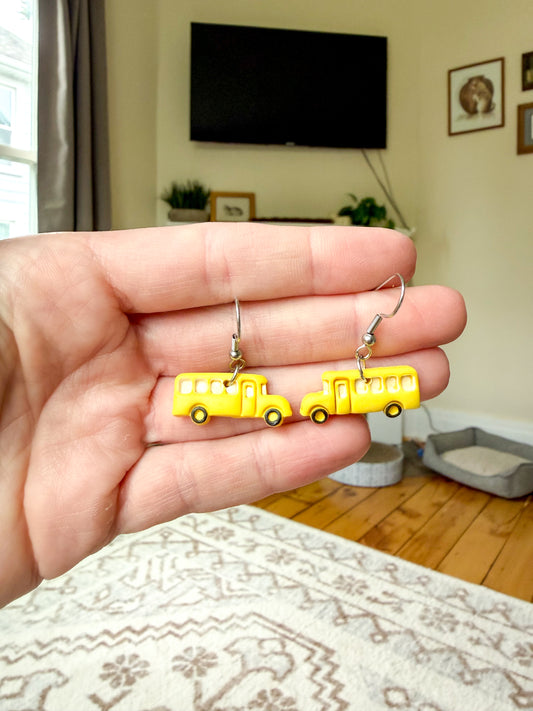 A hand holds a pair of yellow school bus charm earrings with silver fishhook wires, shown against a blurred living room background.