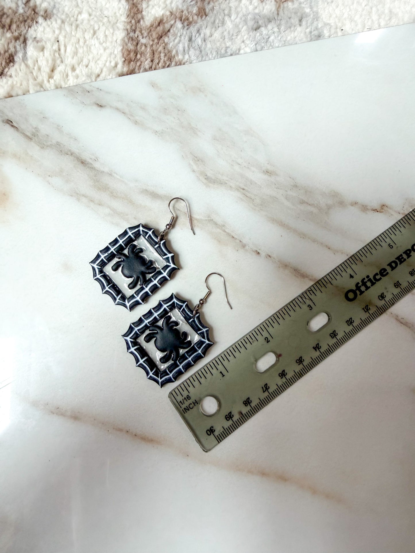 A pair of black and white spiderweb dangle earrings with spider charms, displayed on a white marble surface next to a ruler for scale. Around 2 inches long.