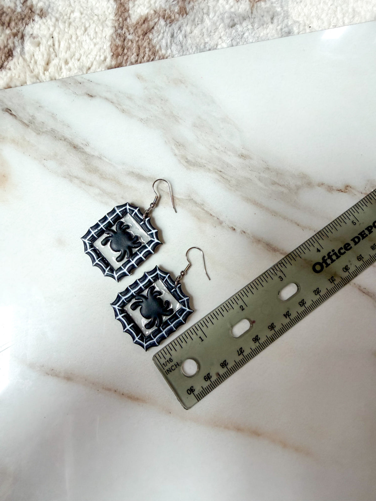 A pair of black and white spiderweb dangle earrings with spider charms, displayed on a white marble surface next to a ruler for scale. Around 2 inches long.