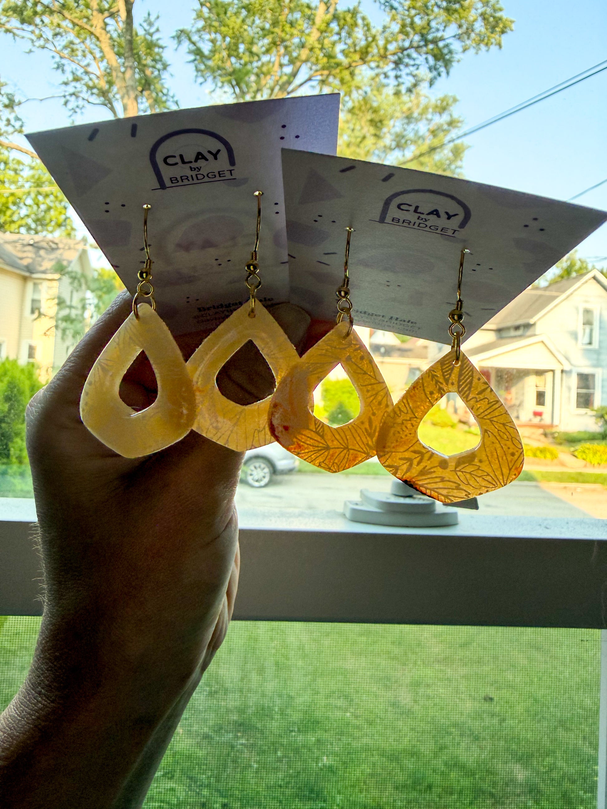 Three pairs of teardrop-shaped dangling earrings displayed on branded cards held by a hand, with a window and outdoor lawn visible in the background. The earrings appear in pale yellow, amber, and golden-yellow tones. They are transluscent in the sun. 