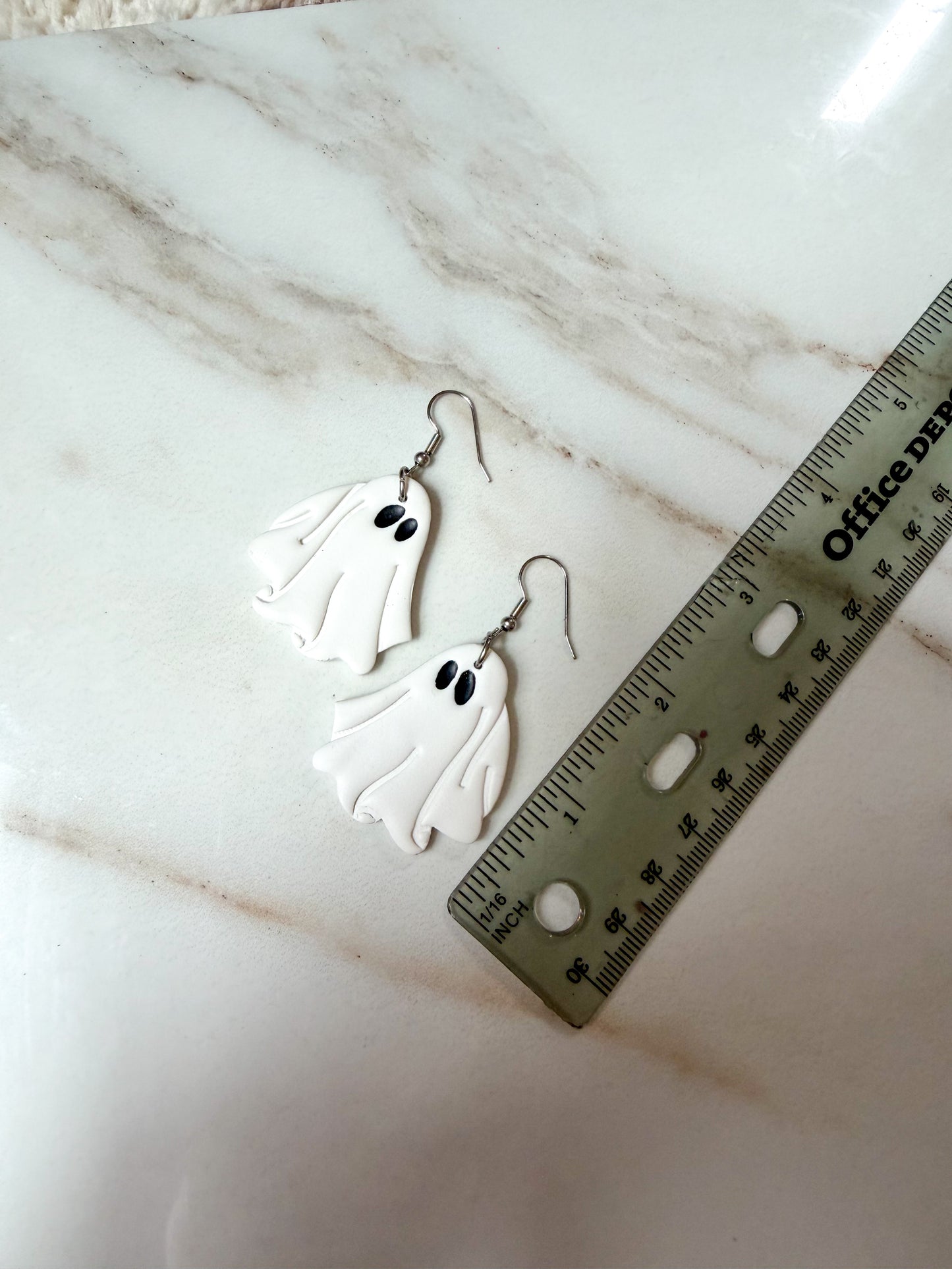 A pair of white ghost-shaped dangle earrings with black eyes and silver hooks, displayed on a white marble surface next to a ruler for scale. Around 2 inches in length. 