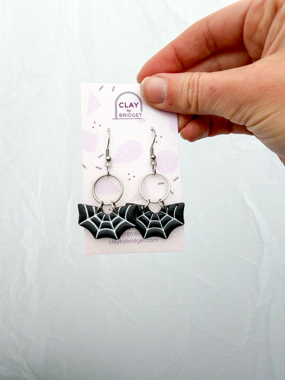 A hand holds a small display card featuring a pair of dangling earrings. The earrings have silver-toned hooks and circular links with black-and-white spiderweb shaped charms.