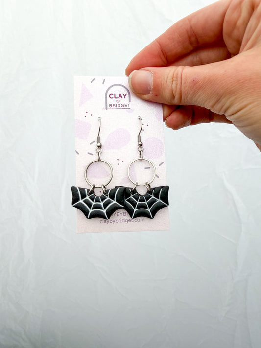 A hand holds a small display card featuring a pair of dangling earrings. The earrings have silver-toned hooks and circular links with black-and-white spiderweb shaped charms.