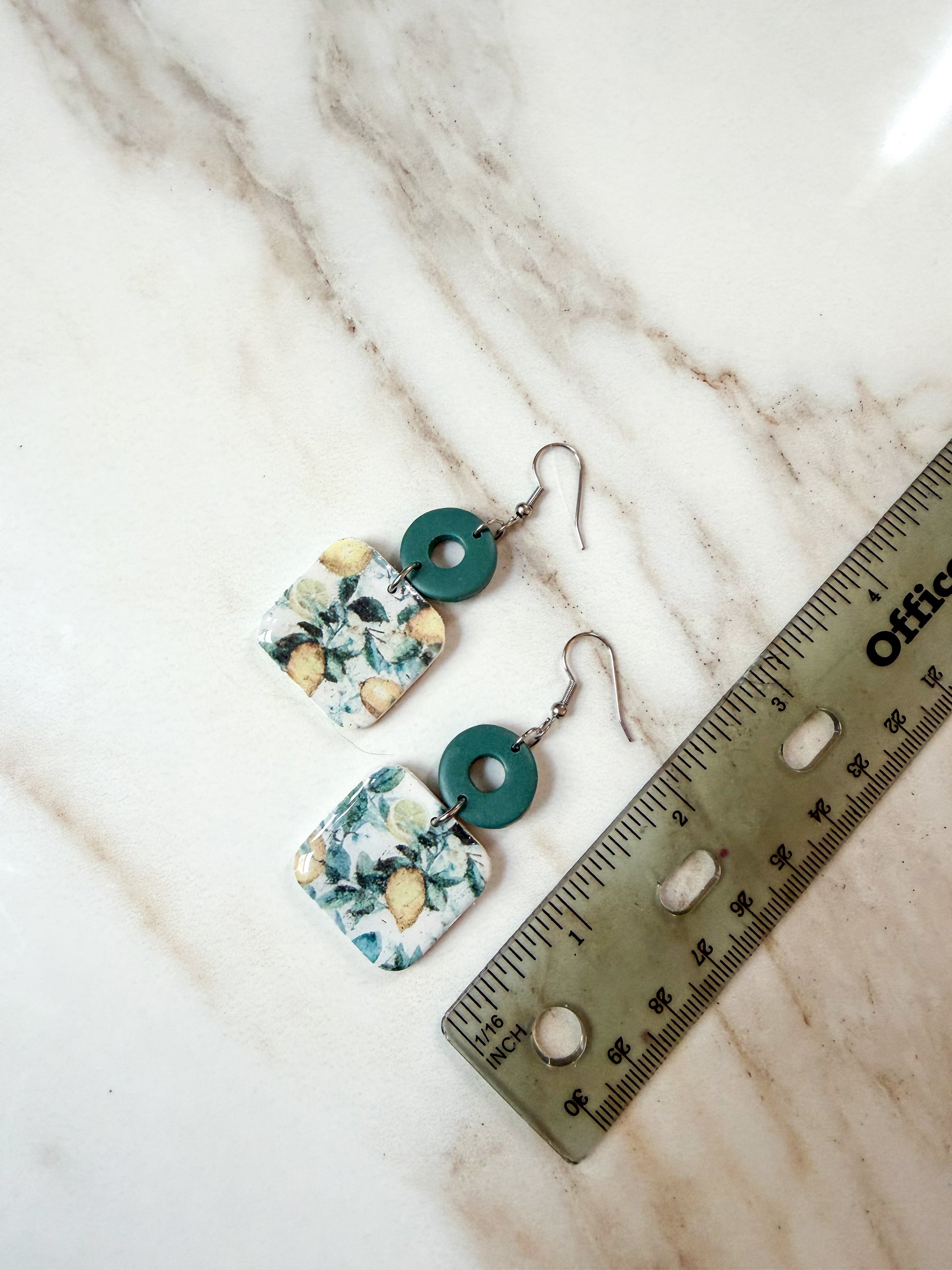 A pair of handmade dangle earrings with square white and green lemon patterned charms, teal green circular accents, and silver fishhook wires, displayed on a white marble surface next to a metal ruler for scale. Nearly 3 inches in length. 